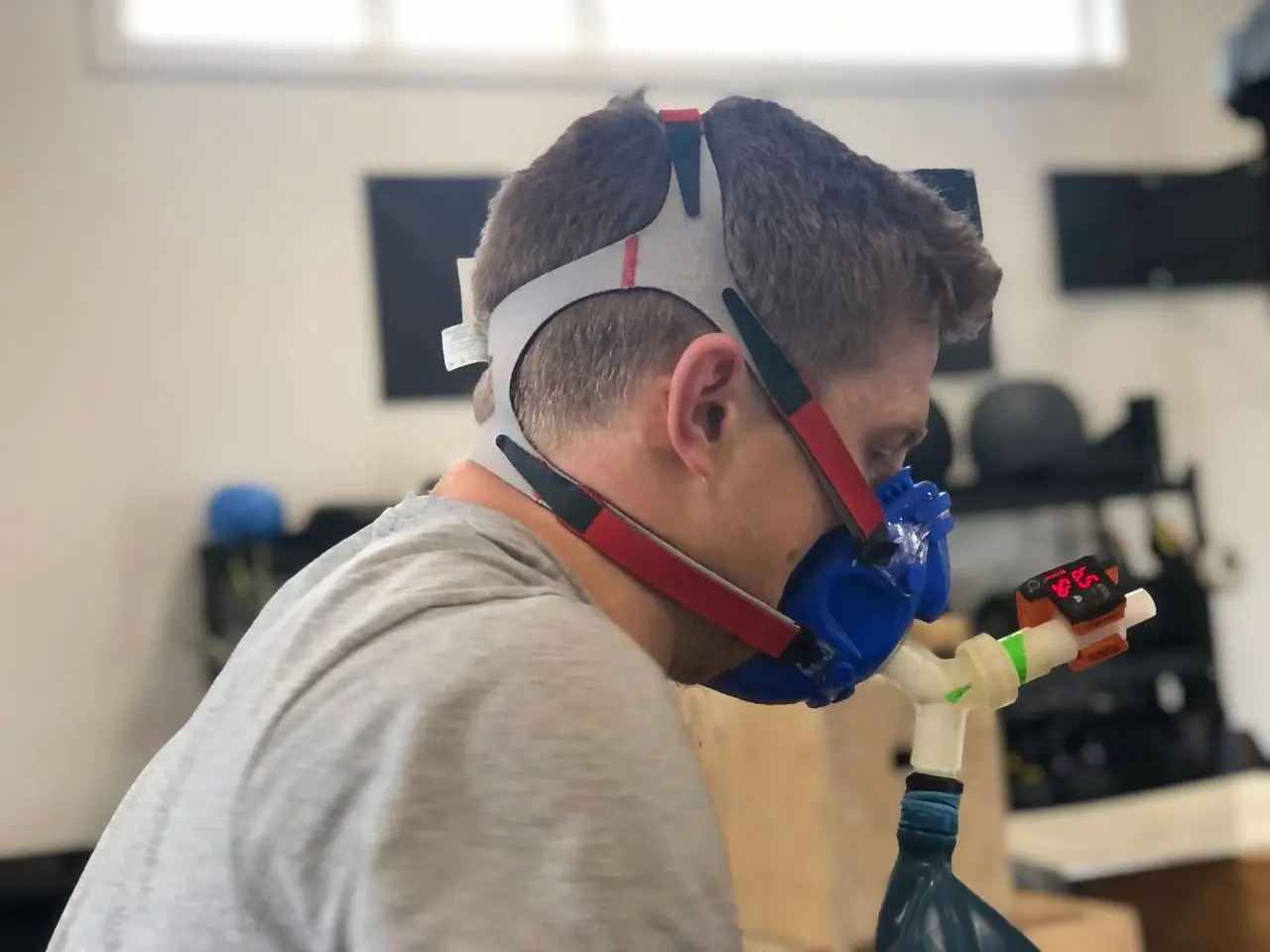 A focused athlete is using the Isocapnic BWB respiratory training device in a high-performance gym setting. The athlete is seated, wearing the device, with their breathing steady and deliberate, simulating a hypercapnic-hypoxic state. Nearby, a coach monitors performance metrics on a tablet, emphasizing precision and progress. In the background, gym equipment and motivational banners hint at preparation for an upcoming competition. The image showcases the athlete’s commitment to improving lung capacity, breathing efficiency, and oxygen utilization while optimizing recovery and raising their anaerobic threshold. This scene highlights how the Isocapnic BWB is revolutionizing breath training for endurance, respiratory endurance, and cardiovascular fitness.